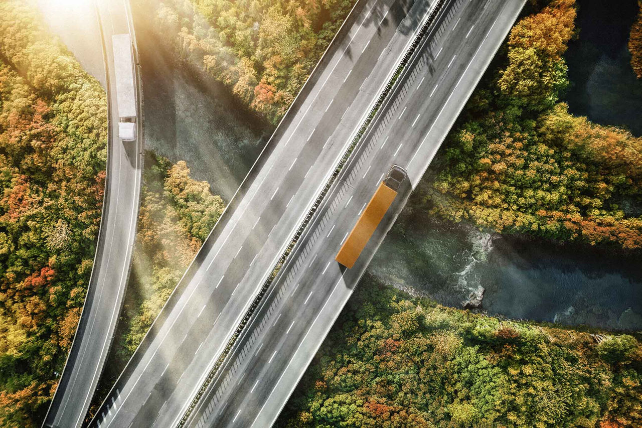 Serviços e Soluções Digitais para Caminhões e Ônibus (Mostra uma vista aérea de uma rodovia que corre sobre um rio sinuoso e cercada por vegetação. Há duas faixas de tráfego visíveis; uma delas, à direita, está ocupada por um caminhão longo de cor predominantemente cinza com uma caçamba laranja. A rodovia apresenta um asfalto claro, e as faixas são demarcadas com linhas brancas. Ao redor, a paisagem é rica em tonalidades de verde, laranja e amarelo, sugerindo que é outono. O rio abaixo é de um tom mais escuro e serpenteia suavemente entre a vegetação, enquanto a luz do sol cria um brilho nas partes superiores da imagem, indicando que é um dia claro)