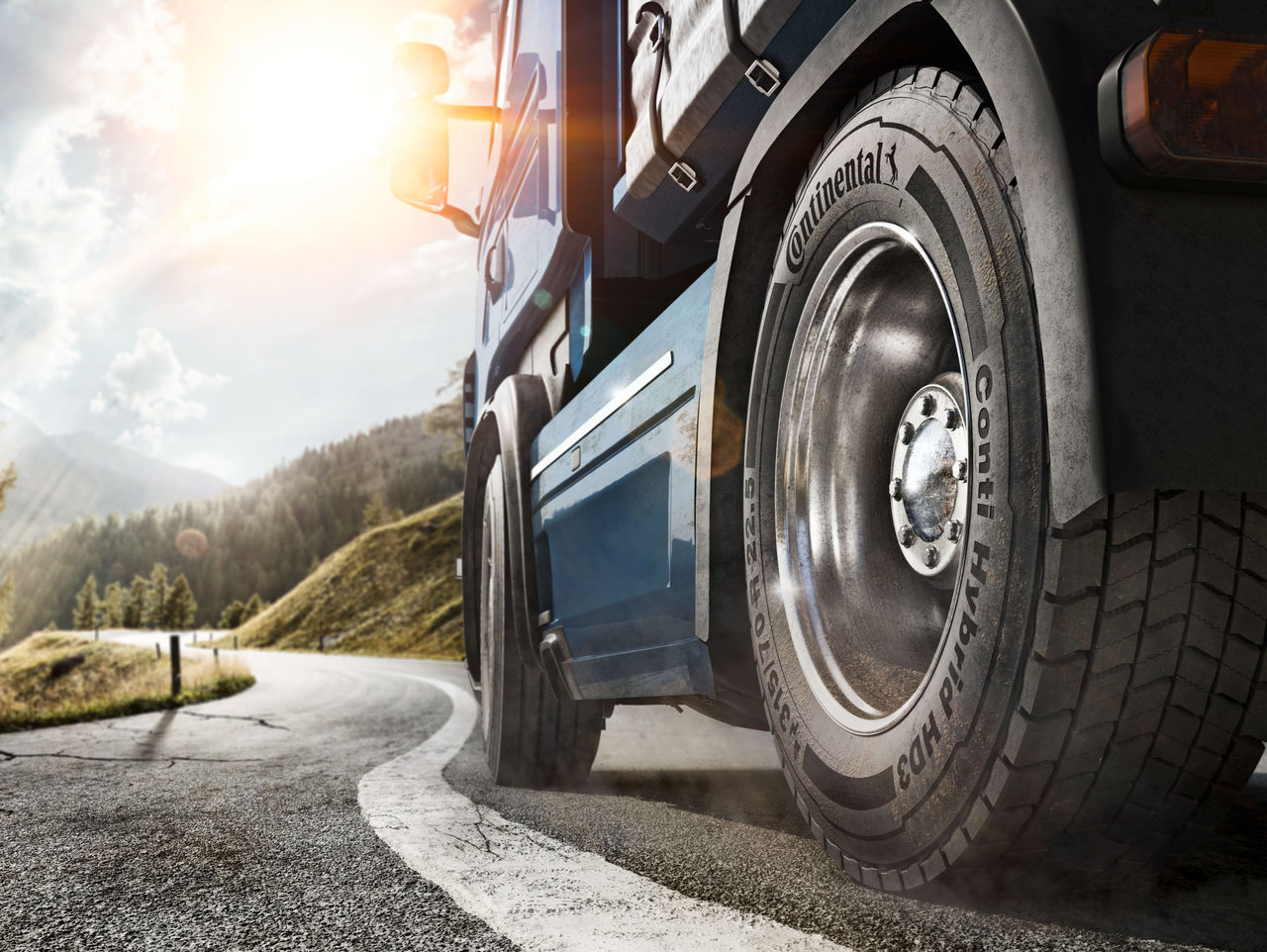 Pneu Continental para Caminhão e Ônibus (A imagem mostra um caminhão grande em uma estrada sinuosa rodeada por uma paisagem montanhosa. A perspectiva é para o lado do caminhão na parte traseira, destacando uma de suas rodas traseiras. O pneu, da marca Continental, é robusto e tem um desenho profundo, indicando que é adequado para estradas, modelo Conti Hybrid HD3. O caminhão em si é pintado de azul e apresenta detalhes prateados, refletindo a luz do sol que vem de um canto da imagem. O céu está parcialmente nublado com raios de sol penetrando as nuvens, criando um ambiente brilhante e natural. Ao fundo, podem-se ver colinas verdes, que adicionam um toque de tranquilidade à cena, enquanto a estrada se curva na direção da imagem)