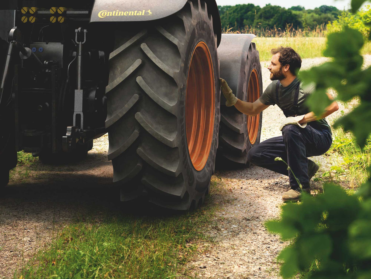 Pneu Continental Agrícola (A imagem retrata um homem agachado ao lado de um trator, inspecionando uma das rodas, que estão com pneu agrícola Continental. Ele usa uma camiseta escura e um par de luvas, e tem cabelos curtos e barba. A roda do trator é grande, com um padrão de sulcos profundos no pneu, a borda da roda é de cor laranja. O cenário é ao ar livre, com um caminho de terra e grama ao redor; ao fundo, há uma vegetação verdejante que sugere um ambiente rural. A luz é natural, indicando um dia ensolarado. A atmosfera transmite uma sensação de trabalho e conexão com a natureza.)