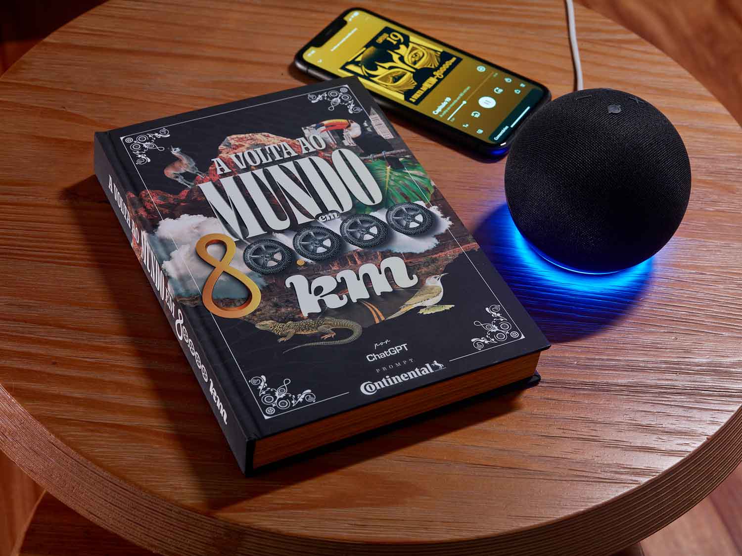 (Livro "A Volta ao Mundo em 80.000 km", inspirado no clássico de Júlio Verne e criado com IA, em uma mesa de madeira ao lado de um dispositivo Alexa com luz azul e um smartphone exibindo o audiolivro no Spotify. Disponível em formatos físico, digital e audiolivro pela Continental Pneus.)