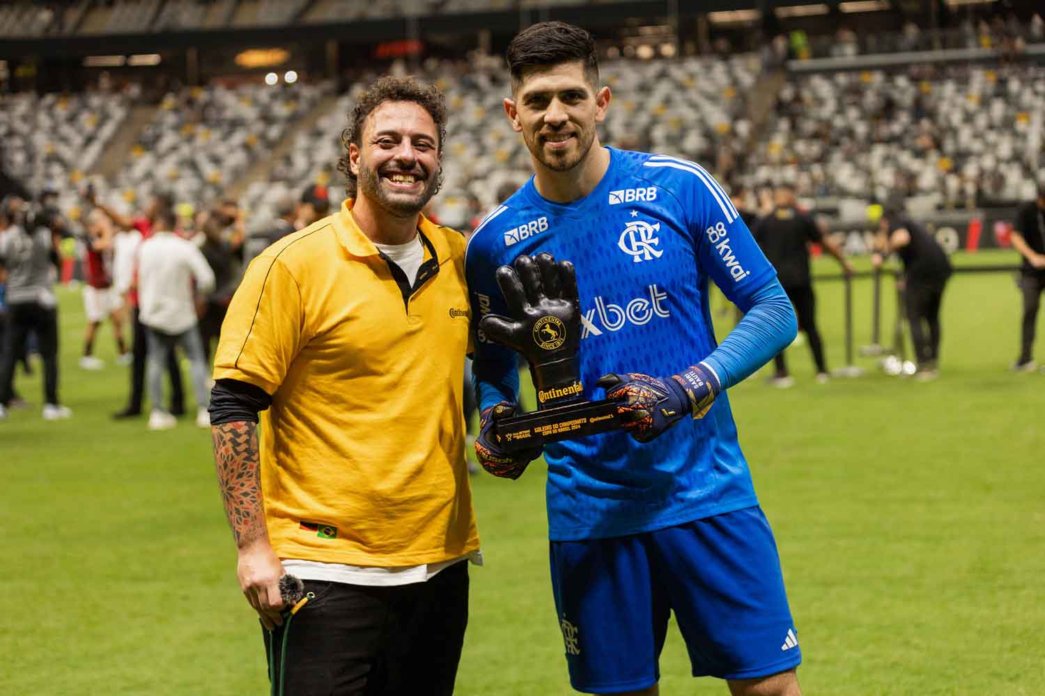 (O influenciador Tomer Savoia, sorrindo e vestindo camisa laranja da Continental, entrega o troféu de melhor goleiro, feito de borracha de pneu Continental, ao goleiro Agustín Rossi que veste uniforme azul do Flamengo e segura o troféu com formato de luva, no campo gramado do estádio, com público ao fundo celebrando a conquista, após a final da Copa do Brasil 2024, onde a Continental é patrocinadora)