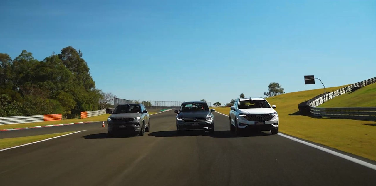 (Imagem de três SUVs lado a lado em movimento em uma pista de corrida durante o dia. Da esquerda para a direita, os veículos são um Jeep Compass cinza escuro, um Volkswagen Tiguan prata e um GWM Haval H6 branco, todos com pneus Continental. Eles aparentam estar em alta velocidade, pois ocupam a largura total da pista, indicando uma cena de ação ou competição. Ao redor da pista, há grama verde e cercas de segurança. No fundo, à direita, há uma colina coberta por árvores e vegetação. Barras de proteção laranjas e brancas são visíveis ao longo dos limites internos da pista, e a pintura da pista inclui faixas brancas e zebras em amarelo e vermelho nas bordas. O ambiente transmite uma sensação de velocidade e esporte a motor. A imagem destaca a comparação de performance entre os modelos Jeep Compass, Volkswagen Tiguan e GWM Haval H6, enfatizando suas características em um ambiente de teste automotivo)