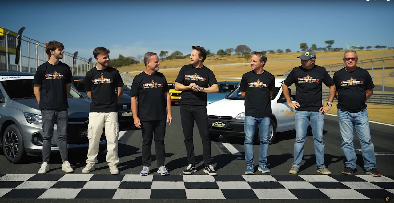 (Grupo de sete pilotos icônicos do automobilismo brasileiro posando na linha de largada/chegada pintada de branco e preto do autódromo Vellocitta, durante a celebração dos 10 anos do canal Acelerados. Eles vestem camisetas pretas com o logo do evento 'Superstars 1.000 by Continental' na frente. Suas expressões são descontraídas e amigáveis, e alguns estão conversando uns com os outros. Ao fundo, diversos carros que serão usados na corrida, incluindo modelos clássicos e modernos, estão alinhados na pista. À esquerda, há um carro de cor cinza e, atrás do grupo, ao centro, um carro amarelo usado como “Safety car” . À direita, há um terceiro veículo, de cor branca. O cenário inclui arquibancadas e áreas gramadas sob um céu azul claro, destacando o ambiente típico de competições automotivas no Brasil. A cena transmite um ambiente casual e alegre, representa uma união de grandes nomes do automobilismo em um evento comemorativo e altamente competitivo)