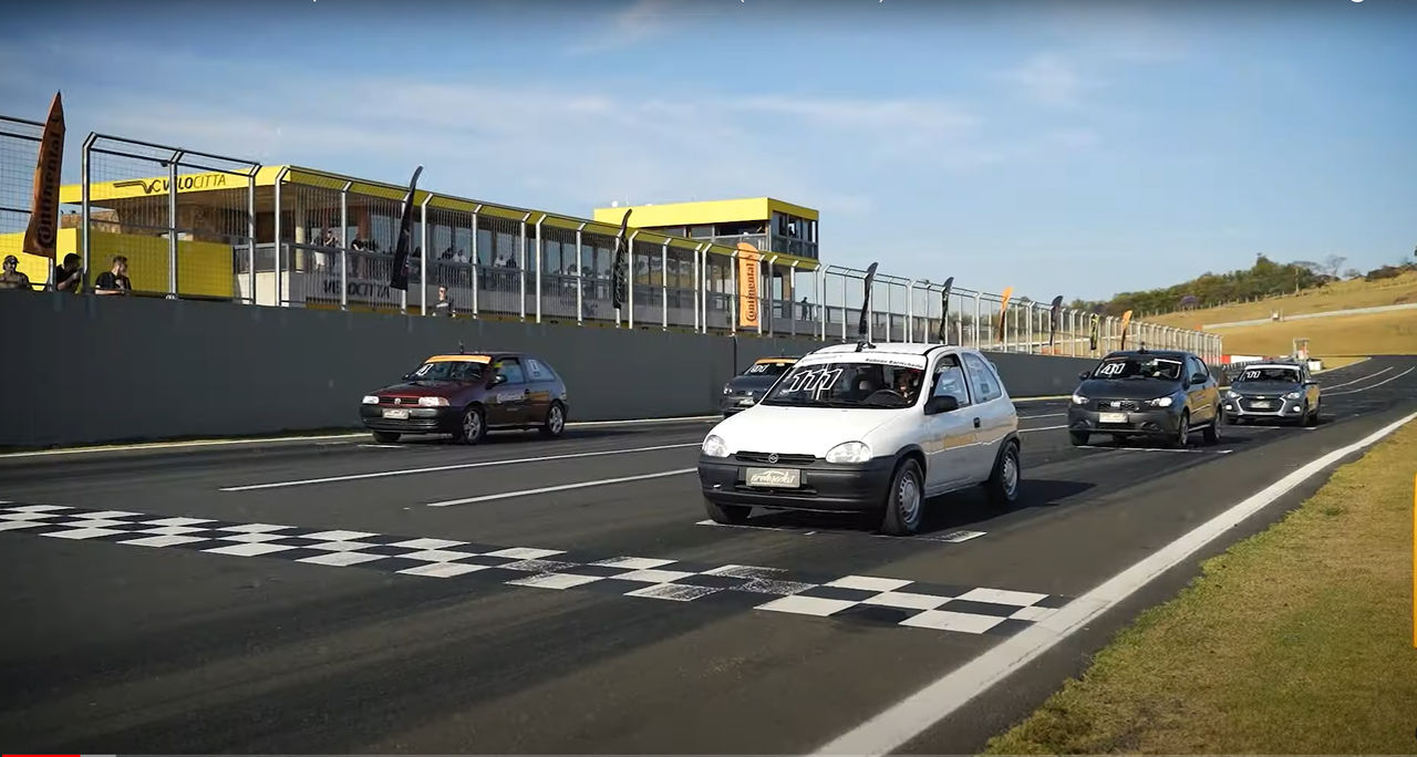 (Esta imagem mostra uma cena de uma corrida de carros populares novos e antigos em um circuito de corridas durante um belo dia com céu claro. Alinhados na linha de largada marcada com padrão quadriculado preto e branco de um autódromo, prontos para a corrida 'Superstars 1.000 by Continental Pneus', comemorativa dos 10 anos do canal Acelerados. Em destaque, um Chevrolet Corsa branco com o número 111 adesivado no para-brisa, ao lado de um Gol vermelho e outros veículos ao fundo, todos equipados com pneus Continental. No alto, acima do muro do circuito, uma arquibancada do autódromo, uma construção amarela com janelas de vidro, está repleta de espectadores. Bandeiras e banners da Continental são visíveis ao fundo acima do muro de proteção ao longo da pista, criando uma atmosfera vibrante e competitiva. A paisagem ao fundo é composta de gramados leves e colinas distantes com vegetação baixa. Esta imagem captura a emoção do início de uma corrida especial, destacando a paixão por carros e a celebração de uma década de conteúdo automotivo)